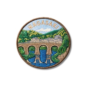 May include: A circular embroidered patch featuring a landscape scene of a stone bridge over blue water with green hills and buildings in the background. The word NAGASAKI is stitched in light tan thread in an arc across the top sky area.