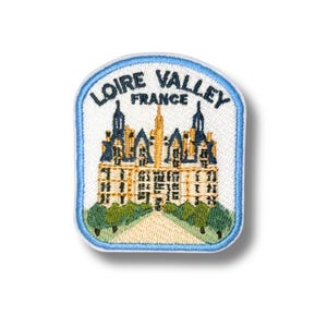 May include: A rectangular embroidered patch with rounded top corners featuring a white background and a light blue border. It depicts a tan castle with blue roofs, green trees, and a path, with the text LOIRE VALLEY FRANCE in dark blue.