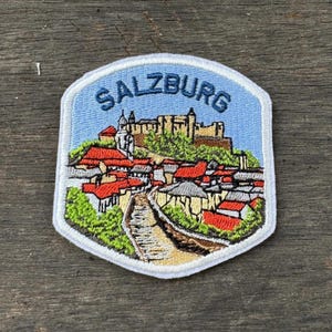 May include: Embroidered patch featuring the word "SALZBURG" above a detailed illustration of a European town. The patch depicts buildings with red roofs, a stone path, and a castle on a hill, all set against a light blue background.