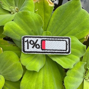 May include: Embroidered patch with a black border, featuring a battery icon showing 1% charge. The battery icon has a red bar indicating low power. The text "1%" is above the battery icon. The patch is set against a background of green leaves.