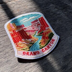 May include: A shield-shaped embroidered patch featuring a landscape of the Grand Canyon with red rock formations, a blue river, and green vegetation. The text GRAND CANYON is stitched in red letters on a white banner at the bottom of the patch.