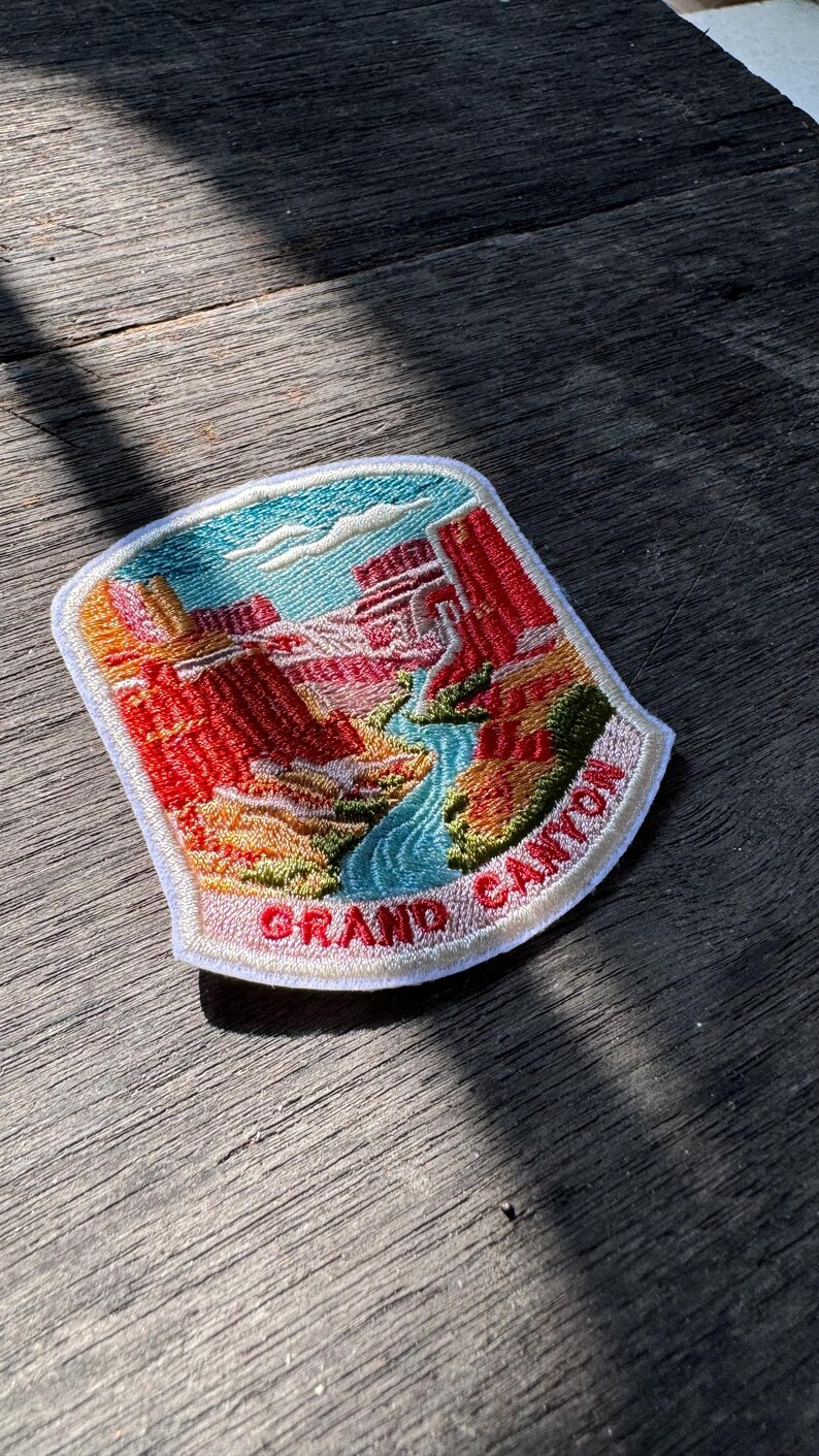 May include: A shield-shaped embroidered patch featuring a landscape of the Grand Canyon with red rock formations, a blue river, and green vegetation. The text GRAND CANYON is stitched in red letters on a white banner at the bottom of the patch.