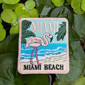 以下が含まれることがあります： 「MIAMI BEACH」の文字が入った、ビーチの風景にピンクのフラミンゴが描かれた刺繍パッチ。パッチには、水色の空、ターコイズブルーの水、緑のヤシの木が描かれています。パッチは正方形で、ピンク色の縁取りがされています。