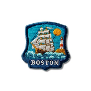 Puede incluir: Un parche bordado en forma de escudo que muestra un velero con velas blancas sobre olas azules, un sol amarillo y un faro rojo y blanco. La palabra BOSTON está bordada en letras mayúsculas blancas en la parte inferior sobre un fondo azul oscuro.