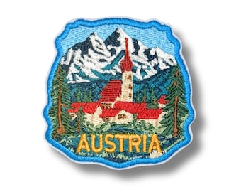 May include: An embroidered patch featuring a mountain landscape with snow-capped peaks, a church with a red roof and steeple, and green pine trees. The word AUSTRIA is stitched in yellow capital letters at the bottom of the blue-bordered design.