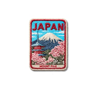 May include: A rectangular embroidered patch featuring a landscape of Mount Fuji, a red pagoda, and pink cherry blossom trees. The word JAPAN is stitched in red at the top, and the text MOUNT FUJI is stitched in red at the bottom on a beige border.