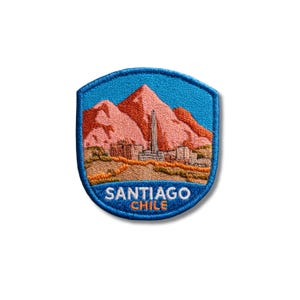 May include: A shield-shaped embroidered patch featuring a landscape of pink mountains behind a city skyline with a tall central tower. The foreground shows brown terrain, and the text SANTIAGO CHILE is stitched in white and orange on a blue border.