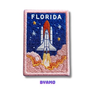 Könnte beinhalten: Gestickter Aufnäher mit einer startenden Raumfähre. Der Aufnäher hat einen blauen Hintergrund mit Sternen und das Wort "FLORIDA" oben. Der Aufnäher ist rosa umrandet und hat das Wort "BYAMO" unten. Die Raumfähre ist weiß und orange.