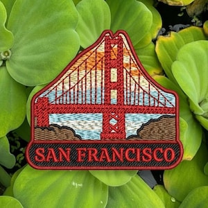 Puede incluir: Parche bordado con el Golden Gate Bridge de San Francisco. El parche muestra un puente rojo sobre un atardecer, agua azul y tierra marrón. El texto "SAN FRANCISCO" está bordado en negro.