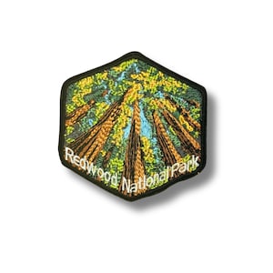 Pode incluir: Um emblema bordado hexagonal com uma vista de ângulo baixo de sequoias altas com troncos castanhos e folhagem verde contra um céu azul claro. O texto Redwood National Park está bordado a branco ao longo da extremidade inferior do emblema.
