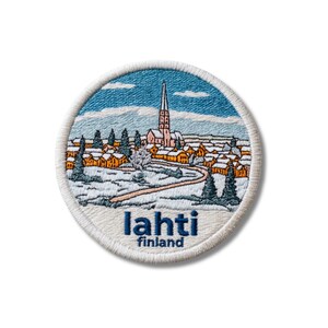 May include: A circular embroidered patch featuring a winter landscape of Lahti, Finland. The design shows a tall church spire, orange-roofed buildings, and snow-covered ground with evergreen trees under a blue sky. The text reads lahti finland.