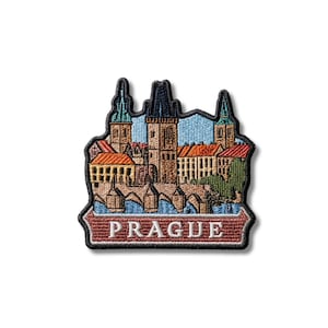 May include: An embroidered patch featuring a detailed cityscape of Prague with orange rooftops, stone towers, and a bridge over blue water. The word PRAGUE is stitched in white capital letters on a dark red banner at the bottom of the patch.