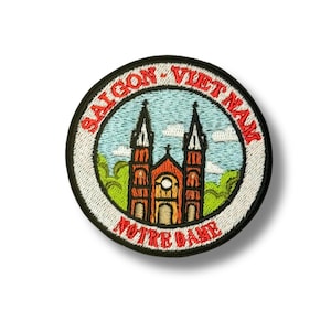 May include: Embroidered patch featuring the Notre Dame Cathedral in Saigon, Vietnam. The design includes the cathedral in shades of orange and brown, set against a blue sky with white clouds. The text "SAIGON - VIETNAM" and "NOTRE DAME" are in red.