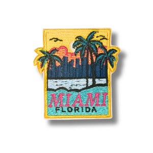 Könnte beinhalten: Ein rechteckiger bestickter Aufnäher mit einer Skyline von Miami, Florida, inklusive Palmen, Sonnenuntergang und Wasser. Der Text MIAMI FLORIDA ist in rosa und schwarzem Garn auf einem gelben Rand und hellblauem Wasserhintergrund gestickt.