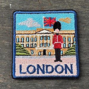 Puede incluir: Parche bordado con una escena de Londres. El diseño incluye el Palacio de Buckingham, una bandera británica y un guardia real con uniforme rojo. La palabra "LONDON" está bordada en letras azules sobre un fondo rosa.