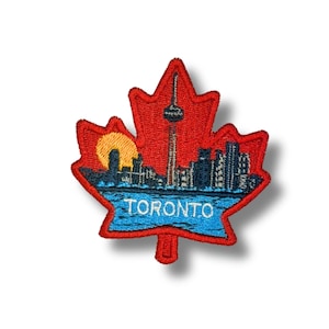 May include: Embroidered patch in the shape of a red maple leaf. The patch features a blue skyline with the text "TORONTO" and a yellow sun. The CN Tower is visible in the center of the skyline.