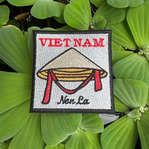 Puede incluir: Un parche blanco bordado con las palabras "VIETNAM" en rojo y "Non La" en cursiva negra. El parche presenta una imagen de dibujos animados de un sombrero cónico vietnamita tradicional.