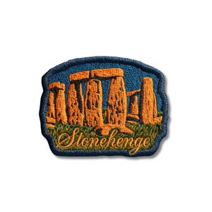 May include: An embroidered patch featuring a depiction of Stonehenge in orange and brown thread against a blue sky background. The base of the monument is surrounded by green grass, and the word Stonehenge is stitched in cursive orange script.