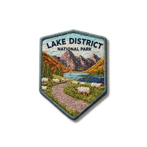 Op de afbeelding: Een schildvormige geborduurde patch met een landschap van het Lake District National Park. Het ontwerp bevat donkerblauwe tekst, bruine bergen, een blauw meer, groene heuvels en een kronkelend pad met diverse witte schapen in het gras.