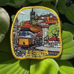 May include: Embroidered patch featuring a Lisbon cityscape with a yellow tram, red-tiled roofs, and the word "LISBON." The patch has a yellow border and is set against a background of green aquatic plants.