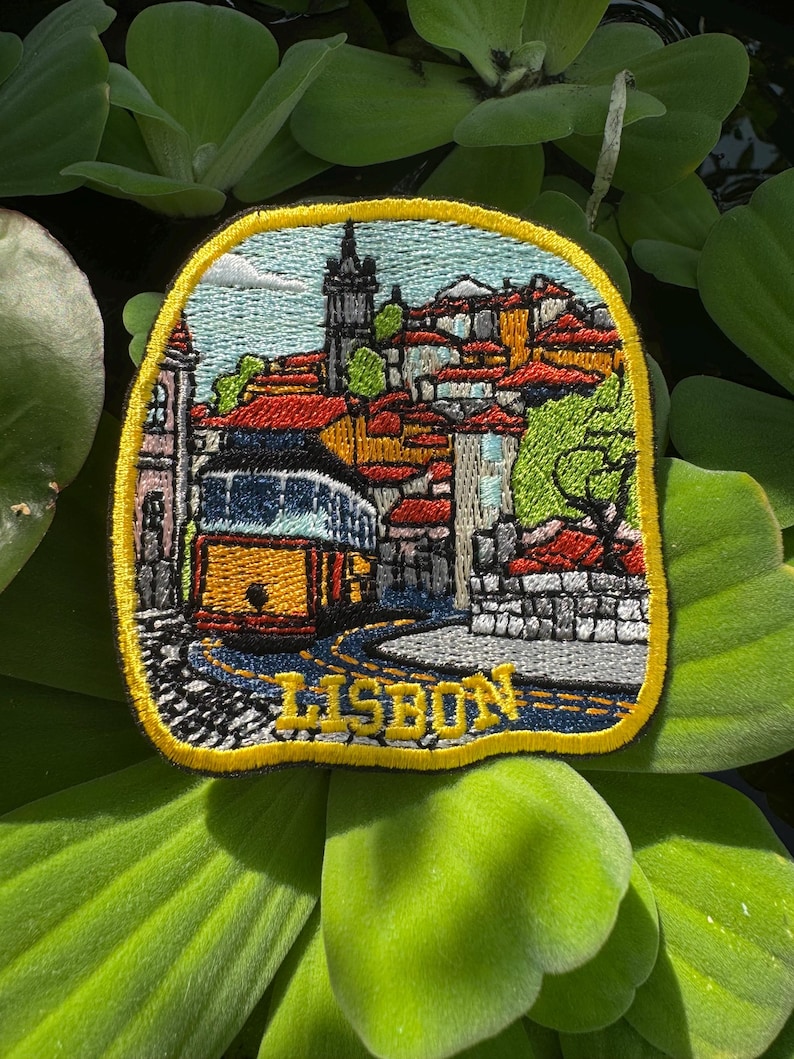 May include: Embroidered patch featuring a Lisbon cityscape with a yellow tram, red-tiled roofs, and the word "LISBON." The patch has a yellow border and is set against a background of green aquatic plants.