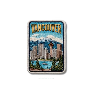 May include: A rectangular embroidered patch featuring the word VANCOUVER in gold letters above a cityscape with a tall tower, blue mountains, and a body of water with a small boat, all framed by green pine trees and a white stitched border.