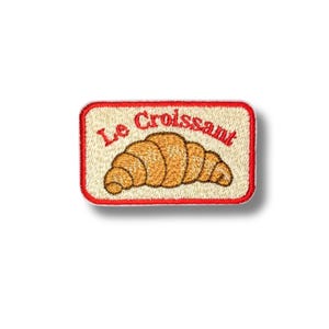 May include: A rectangular embroidered patch with a cream-colored background and a red border. The patch features a golden-brown croissant illustration centered above the red embroidered text that reads Le Croissant in a cursive script font.