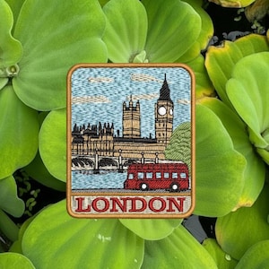 May include: Embroidered patch featuring a London scene with Big Ben, the Houses of Parliament, a red double-decker bus, and the word "LONDON" in a gold border. The patch is set against a background of green aquatic plants.