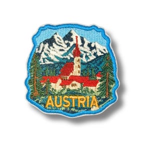 May include: Embroidered patch depicting an Austrian landscape. It shows a church with a red roof, mountains with snow, and fir trees, enclosed within a blue border. The word "AUSTRIA" is embroidered in gold.