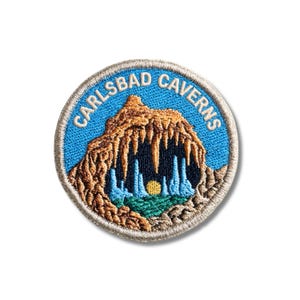 Può includere: Una toppa ricamata circolare con uno sfondo di cielo blu e la scritta CARLSBAD CAVERNS in bianco. Il centro raffigura l'ingresso di una grotta rocciosa marrone con stalattiti color cuoio, stalagmiti azzurre, un sole giallo e terreno verde.