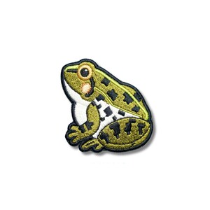 May include: An embroidered iron-on patch shaped like a frog. The frog is olive green with black spots, a white belly, and a large brown eye. The entire design is outlined in a thick black border, creating a distinct and clear graphic shape.