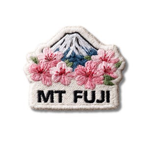 Può includere: Una toppa ricamata che raffigura il monte Fuji in bianco e blu, circondato da fiori di ciliegio rosa e foglie verdi. Il testo MT FUJI è ricamato in grassetto nero su uno sfondo bianco alla base del disegno.