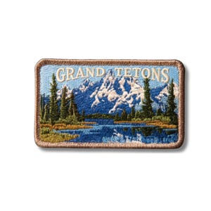 May include: A rectangular embroidered patch featuring a landscape of the Grand Tetons with snow-capped mountains, a blue lake, and green pine trees. The text GRAND TETONS is stitched in white at the top, and the patch has a brown stitched border.