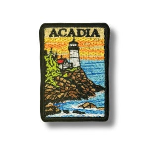 May include: Embroidered patch featuring a lighthouse on a rocky coast with the word "ACADIA" in gold lettering. The design includes a sunset sky, blue water, and green foliage, all framed by a black border.