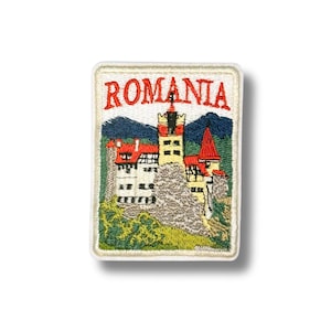 May include: A rectangular embroidered patch featuring the word ROMANIA in red capital letters at the top. The design depicts a castle with white walls, red roofs, and a tall tower, set against a backdrop of dark blue mountains and green trees.