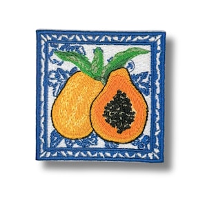 Puede incluir: Un parche bordado cuadrado que presenta una papaya amarilla entera y una mitad de papaya naranja con semillas negras. La fruta está sobre un fondo blanco con patrones florales azules, todo encerrado dentro de un marco de borde azul.