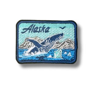 May include: A rectangular embroidered patch featuring a dark blue whale breaching from light blue water. The background shows grey mountains and a small bird. The word Alaska is stitched in dark blue script at the top of the patch.