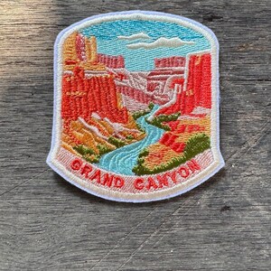 May include: An embroidered patch featuring a landscape of the Grand Canyon with red rock formations, a winding blue river, and a light blue sky with white clouds. The text GRAND CANYON is stitched in red at the bottom on a light tan border.