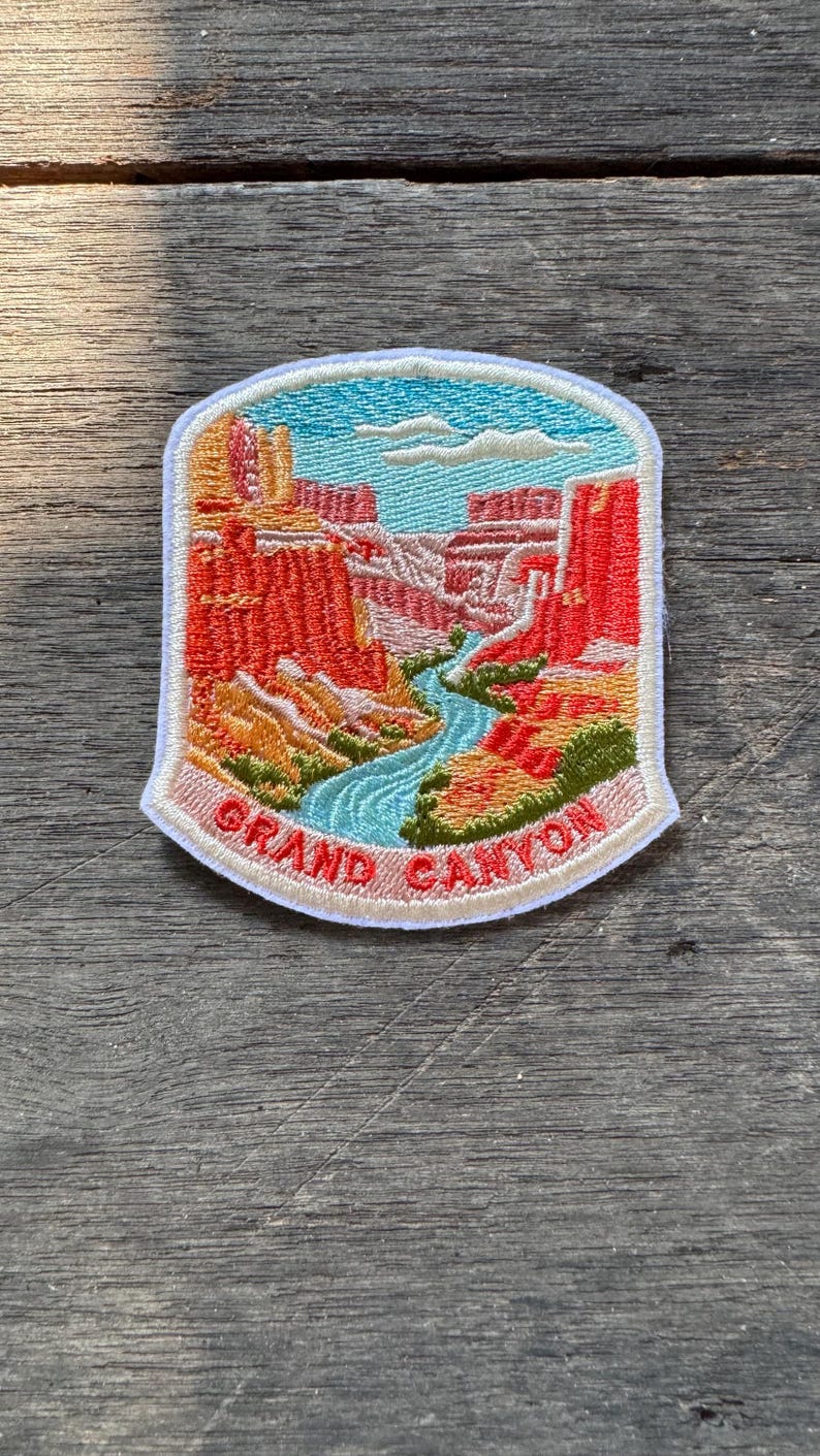 May include: An embroidered patch featuring a landscape of the Grand Canyon with red rock formations, a winding blue river, and a light blue sky with white clouds. The text GRAND CANYON is stitched in red at the bottom on a light tan border.