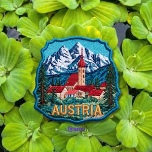 May include: Embroidered patch featuring a scenic Austrian landscape with snow-capped mountains, a church with a red roof and steeple, and the word "AUSTRIA". The patch has a blue border and is set against a background of green aquatic plants.