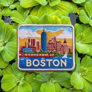 Puede incluir: Parche bordado con el horizonte de Boston, con un fondo de agua y cielo azules. El parche lleva la palabra "BOSTON" en letras azules. El parche está enmarcado por un borde blanco y sobre un fondo de plantas acuáticas verdes.