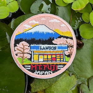 May include: Embroidered patch featuring Mt. Fuji with a blue and white mountain, a Lawson convenience store, and cherry blossom trees. The patch has the text "LAWSON" and "MT. FUJI JAPAN" in a circular design with a pink border.
