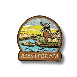 May include: Embroidered patch featuring an Amsterdam scene. The design includes a windmill, canal, boat, and buildings under a blue and yellow sky. The word "AMSTERDAM" is embroidered at the bottom in a brown border.