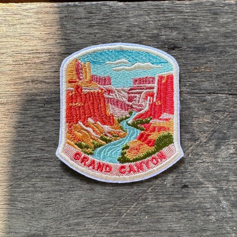 May include: A shield-shaped embroidered patch featuring a landscape of the Grand Canyon with red rock formations, a winding blue river, and white clouds in a light blue sky. The text GRAND CANYON is stitched in red at the bottom of the patch.