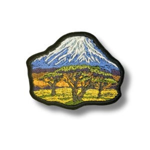 May include: Embroidered patch featuring a mountain with a snow-capped peak, blue sky, and green and yellow trees. The patch has a black border and depicts a landscape scene.