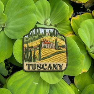 Puede incluir: Parche bordado con un paisaje toscano con colinas, cipreses y una villa. El parche tiene forma rectangular con esquinas redondeadas y la palabra "TUSCANY" en una fuente serif.