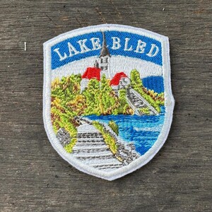 Può includere: Toppa ricamata raffigurante il lago di Bled. La toppa ha un bordo bianco e una parte superiore blu ad arco con le parole "LAKE BLED". L'immagine raffigura una chiesa con tetti rossi, alberi verdi, un lago e un ponte di pietra.