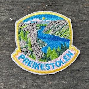 May include: Embroidered patch featuring a scenic landscape with a river, mountains, and trees. The patch has a blue sky with a sun and the text "PREIKESTOLEN" in pink. The patch is bordered with yellow and white.
