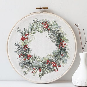 May include: A green and white Christmas wreath with red berries, embroidered on a white fabric and displayed in a wooden hoop.