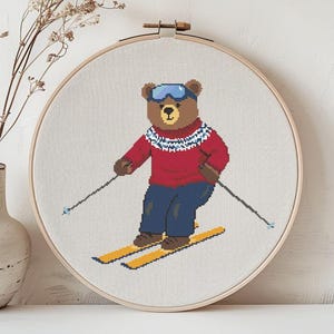 May include: Cross-stitch embroidery of a bear skiing, wearing a red sweater, blue pants, and goggles. The bear is on yellow skis, holding ski poles. The embroidery is in a wooden hoop, with a vase of dried flowers to the left.
